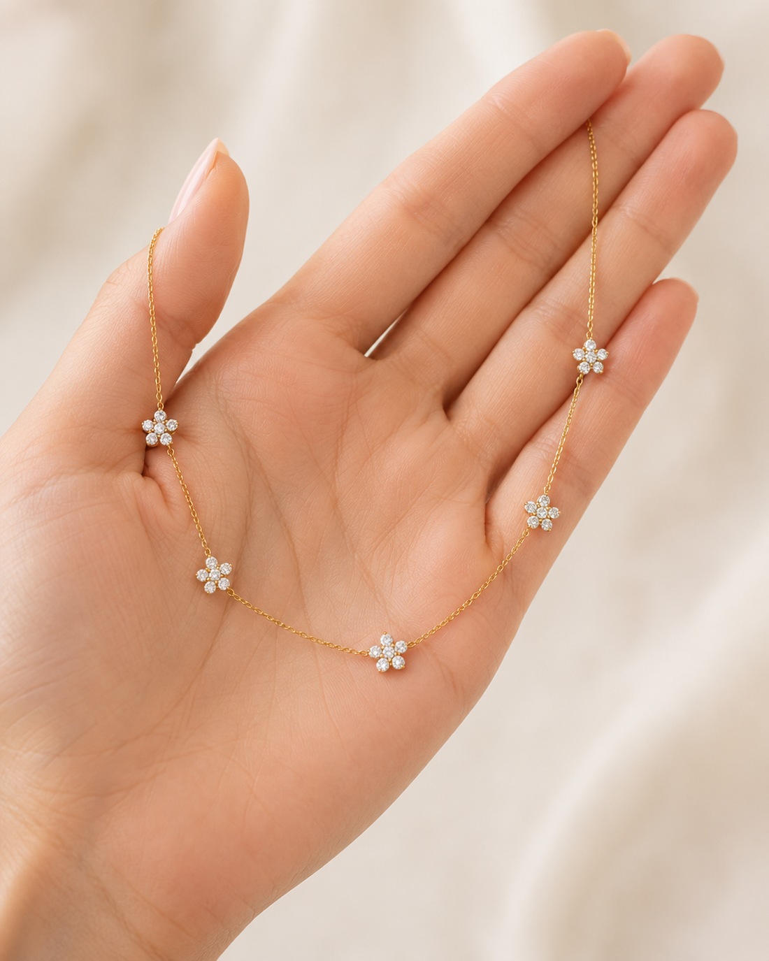 A delicate gold chain necklace with five small white flower charms, held in a person's hand against a soft, light beige fabric background.