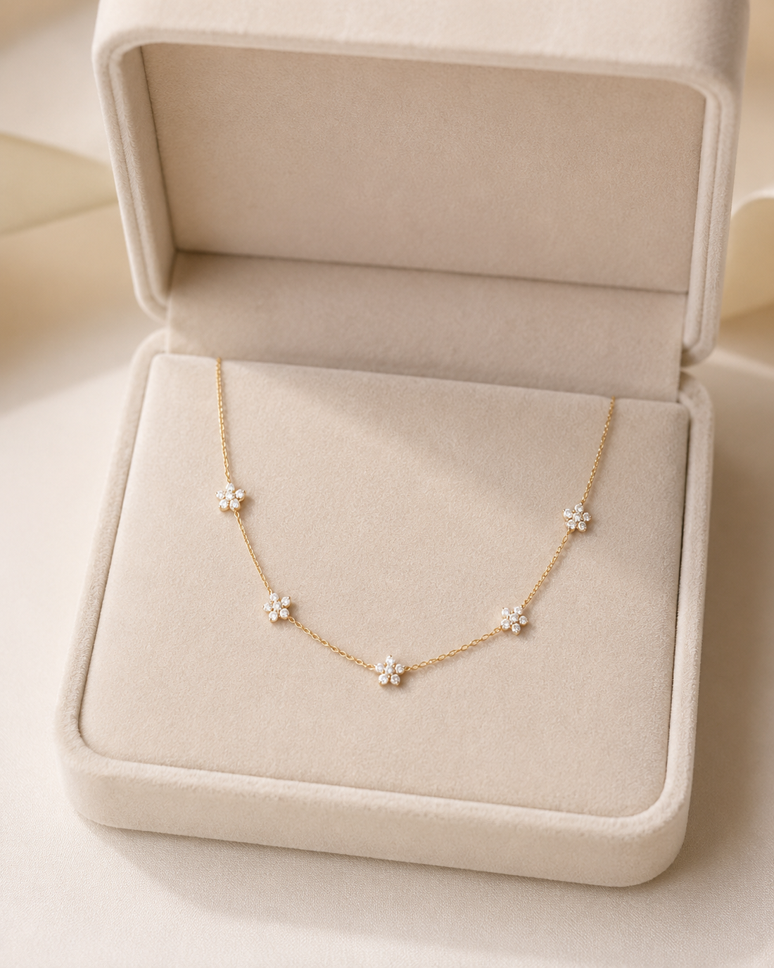 A delicate gold necklace with five small flower clusters, each set with clear stones, displayed inside an open cream-colored jewelry box.