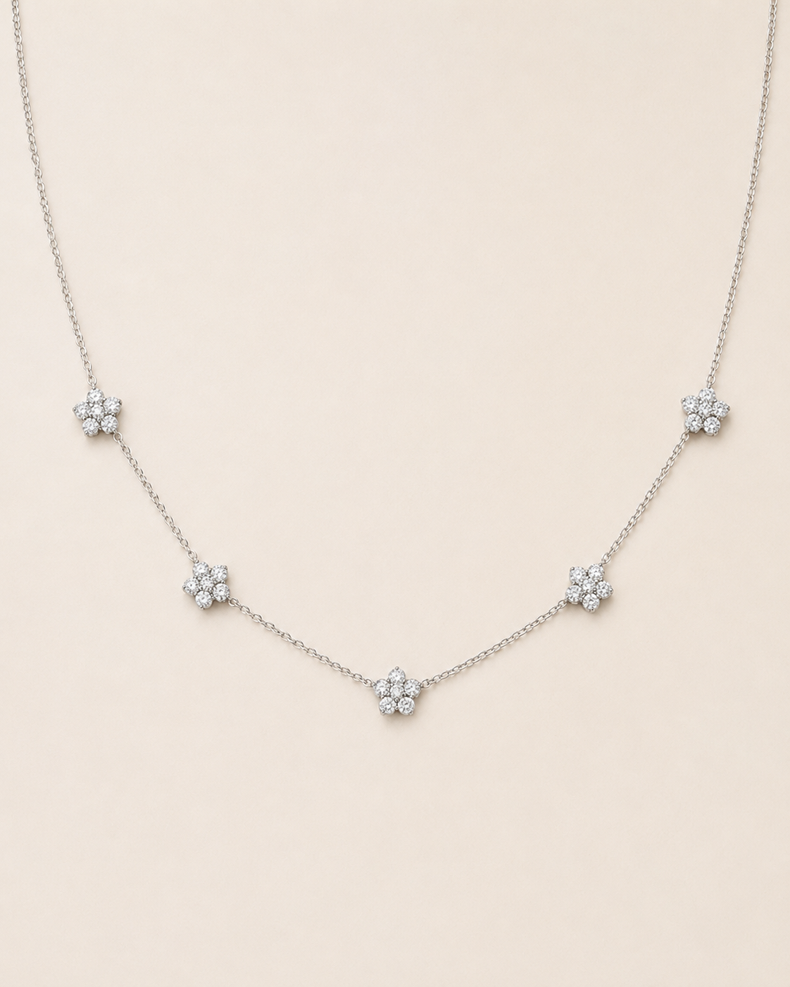 A delicate silver chain necklace featuring five small, flower-shaped clusters of clear stones spaced evenly along the chain, displayed against a solid light beige background.
