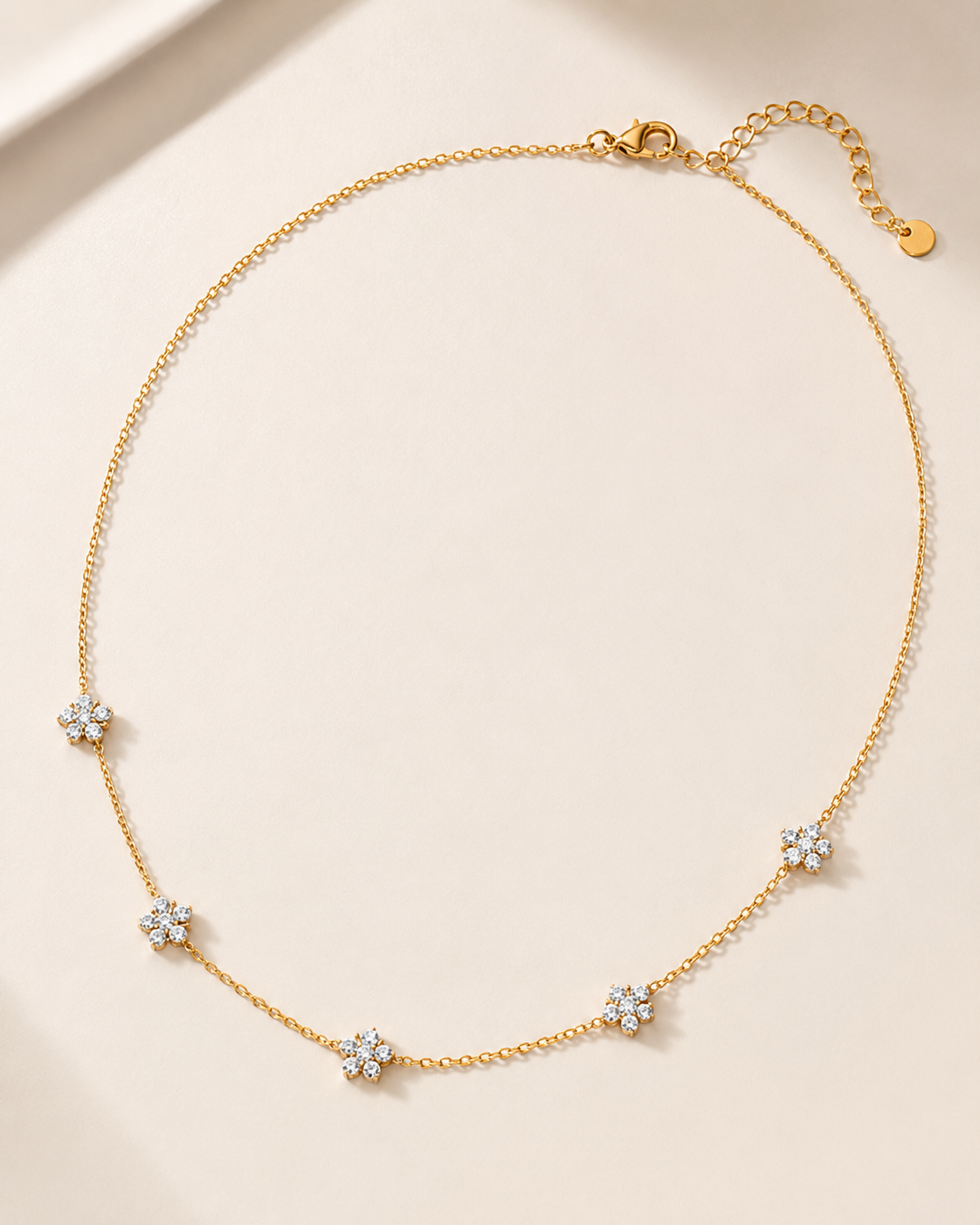 A delicate gold chain necklace with five small flower-shaped crystal clusters spaced along the chain, displayed on a soft beige background.