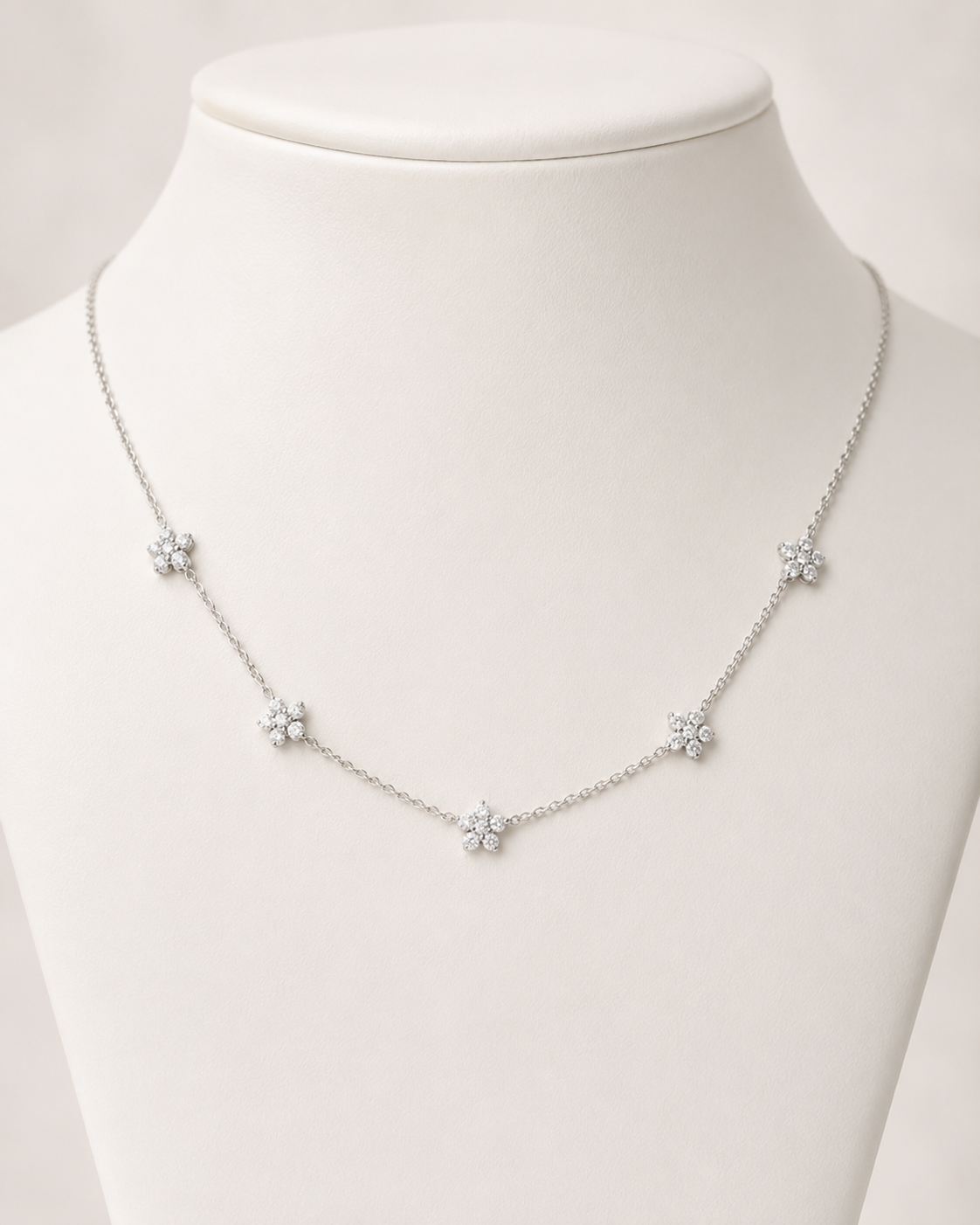 A delicate silver-toned necklace with five small flower-shaped charms, displayed on a white jewelry bust against a soft light background.