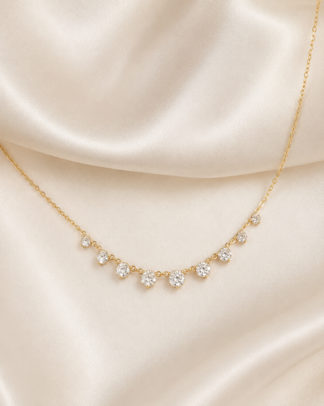 A delicate gold necklace with seven small clear gemstones arranged in a gentle arc, displayed on draped cream satin fabric.