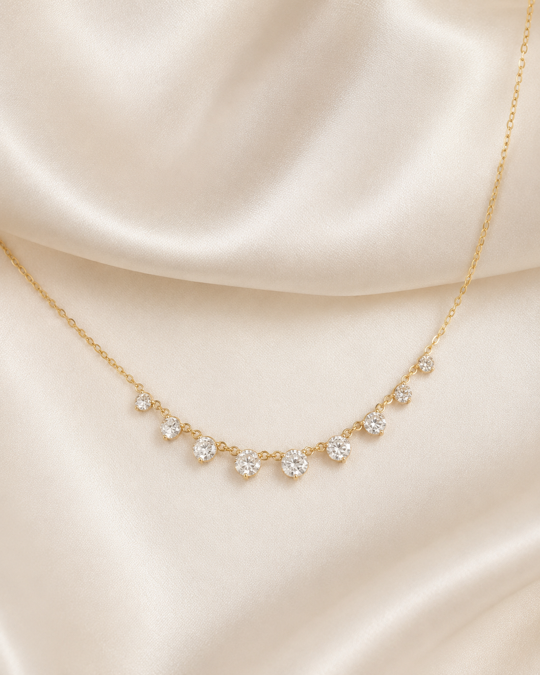 A delicate gold necklace with seven small clear gemstones arranged in a gentle arc, displayed on draped cream satin fabric.