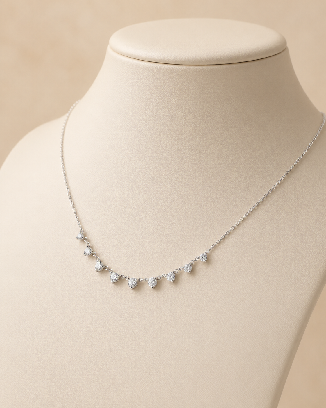 A delicate silver necklace with five small clear teardrop-shaped crystals, displayed on a cream-colored jewelry bust against a soft beige background.