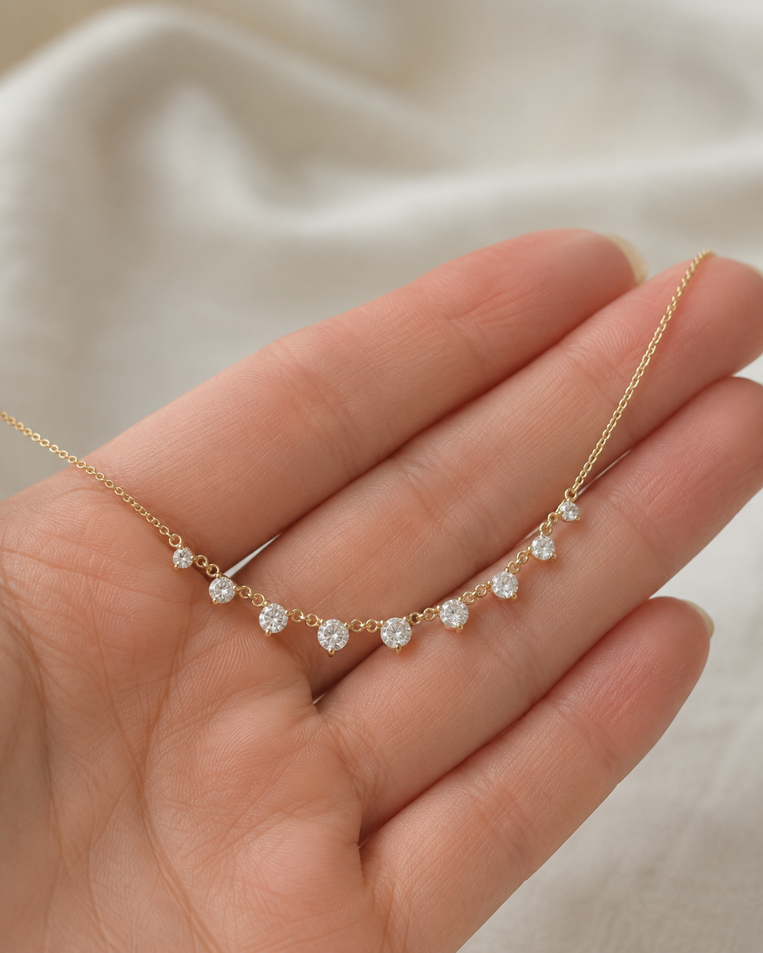 A delicate gold necklace with five small clear gemstones arranged in a gentle arc, held in the palm of a hand against a soft, light beige fabric background.