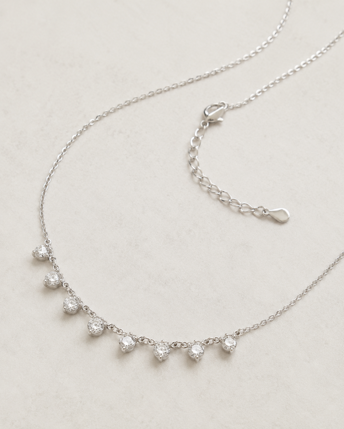 A delicate silver chain necklace with five small clear gemstones set in a curved row, displayed on a light beige textured background. The necklace includes an adjustable lobster clasp extension chain.