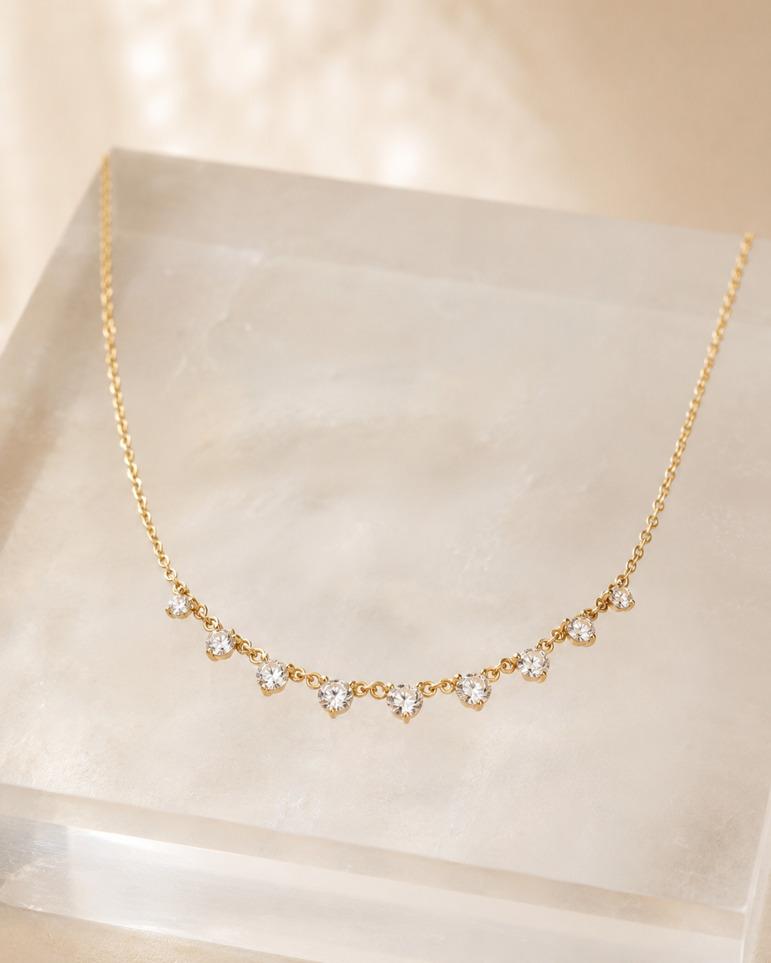 A delicate gold chain necklace with a row of small clear heart-shaped gemstones, displayed on a translucent cream-colored acrylic block against a warm beige background.