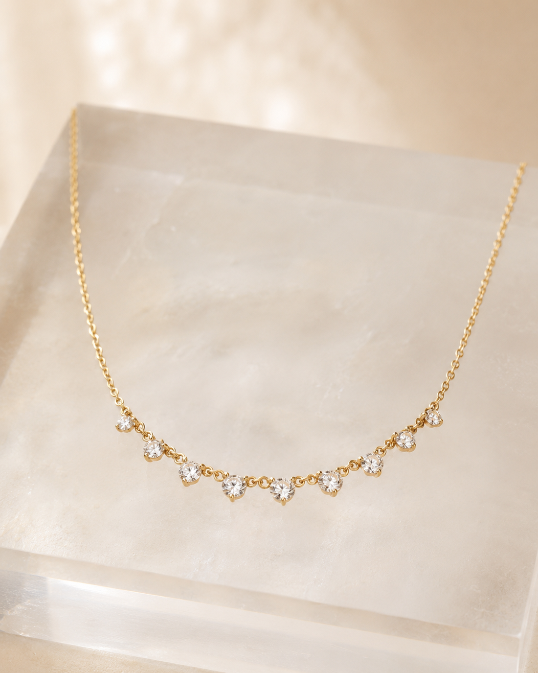 A delicate gold chain necklace with a row of small clear heart-shaped gemstones, displayed on a translucent cream-colored acrylic block against a warm beige background.