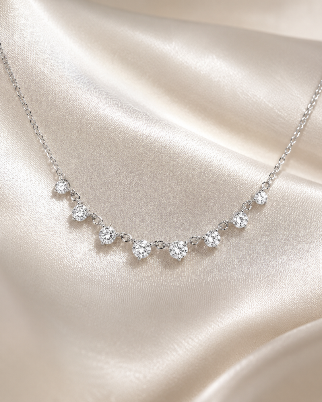 A delicate silver necklace with seven small clear heart-shaped gemstones arranged in a row, displayed on draped champagne-colored satin fabric.