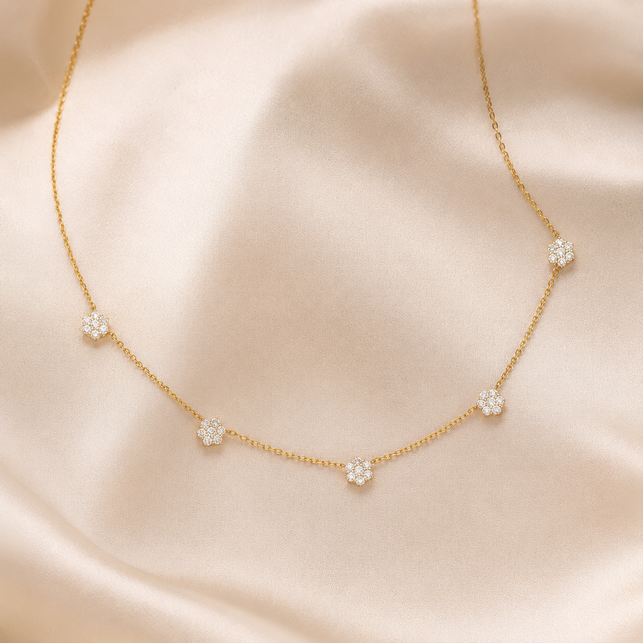 A delicate gold chain necklace with five small flower-shaped clusters of clear stones, displayed on a soft beige satin fabric background.