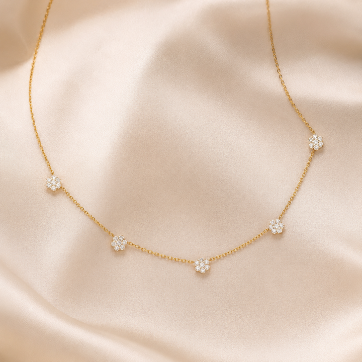 A delicate gold chain necklace with five small flower-shaped clusters of clear stones, displayed on a soft beige satin fabric background.