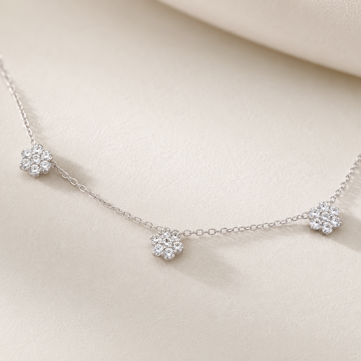 A delicate silver chain necklace featuring three small, round cluster floral charms set with clear stones, displayed on a soft beige fabric background.