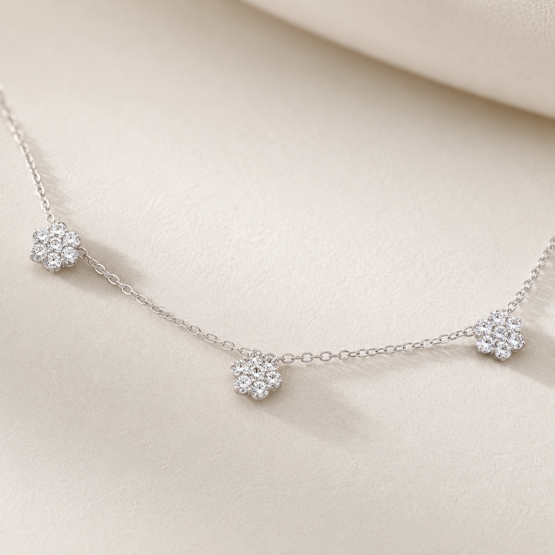 A delicate silver chain necklace featuring three small, round cluster floral charms set with clear stones, displayed on a soft beige fabric background.
