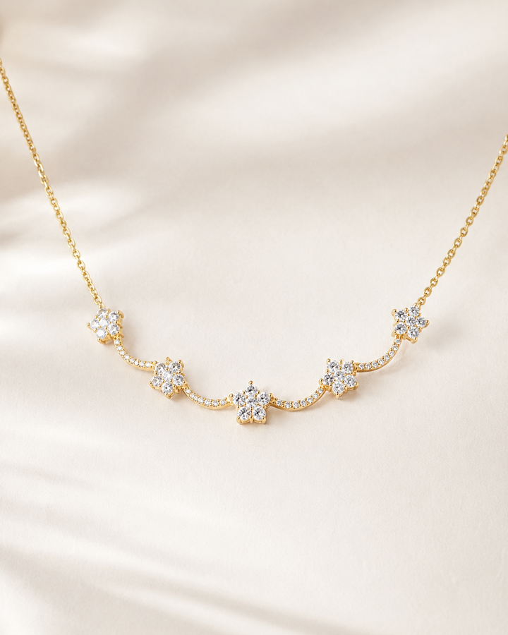 A delicate gold chain necklace featuring five small flower-shaped clusters of clear stones, displayed on a soft, light beige satin fabric background.