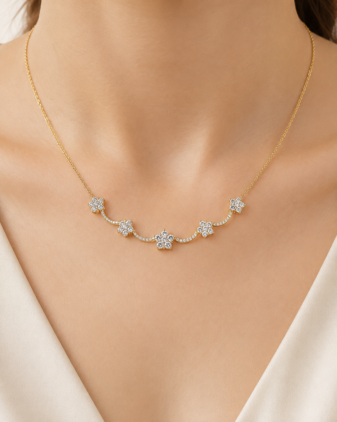 A delicate gold necklace with five small flower-shaped clusters of clear stones, worn on a model's neck over a cream-colored top.