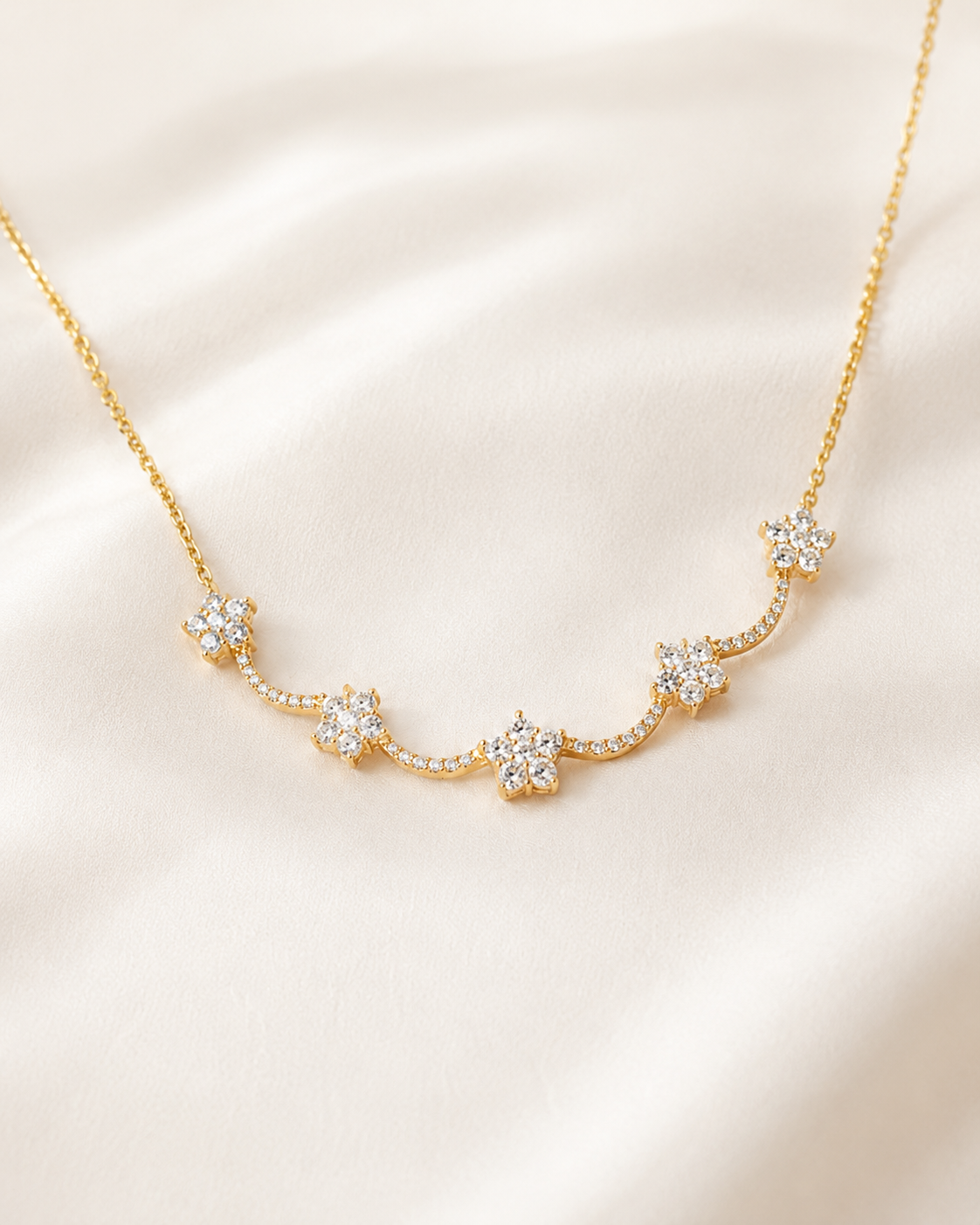 A delicate gold necklace with five small star-shaped clusters of clear stones, arranged in a curved line on a soft, light beige satin fabric background.