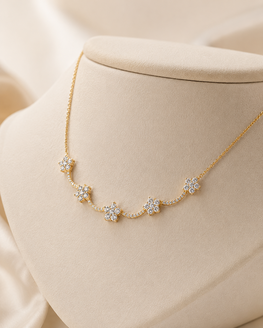 A delicate gold chain necklace with five small flower-shaped clusters of clear stones, displayed on a cream-colored jewelry bust against a soft beige fabric background.