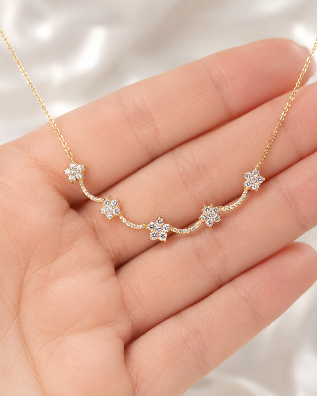 A delicate gold necklace with five small flower-shaped clusters of clear stones, held in the center of a person's hand against a soft white satin background.
