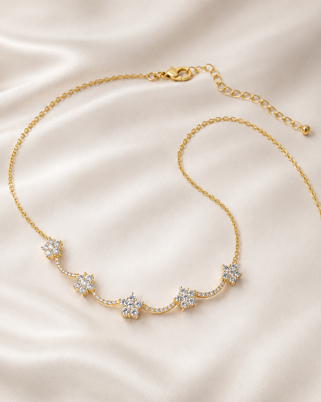 A delicate gold chain necklace with five small star-shaped clusters of clear stones, displayed on a soft beige satin fabric background.