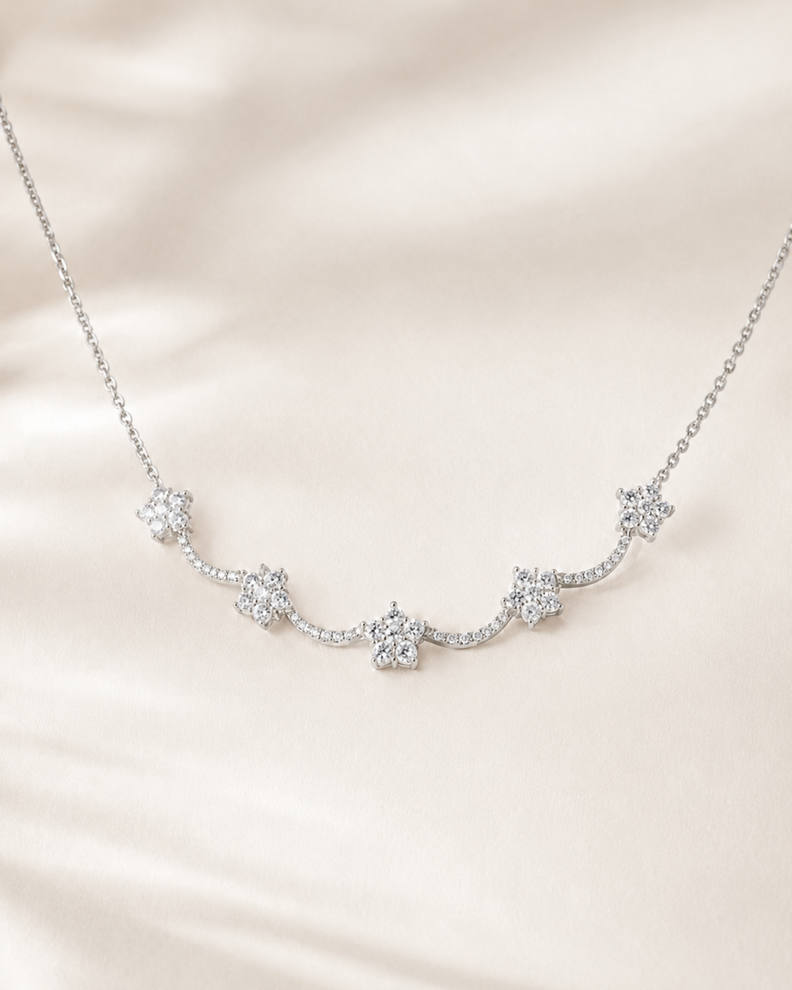 A delicate silver-toned necklace featuring five small flower-shaped clusters of clear stones, displayed on a soft, draped cream satin fabric.