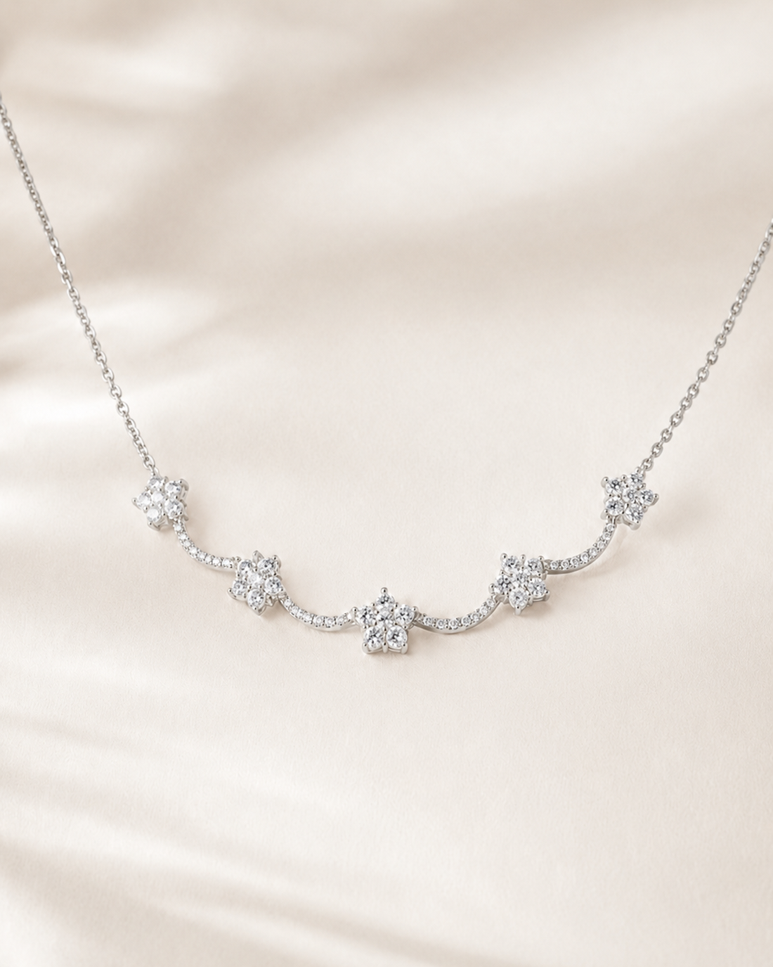 A delicate silver-toned necklace featuring five small flower-shaped clusters of clear stones, displayed on a soft, draped cream satin fabric.