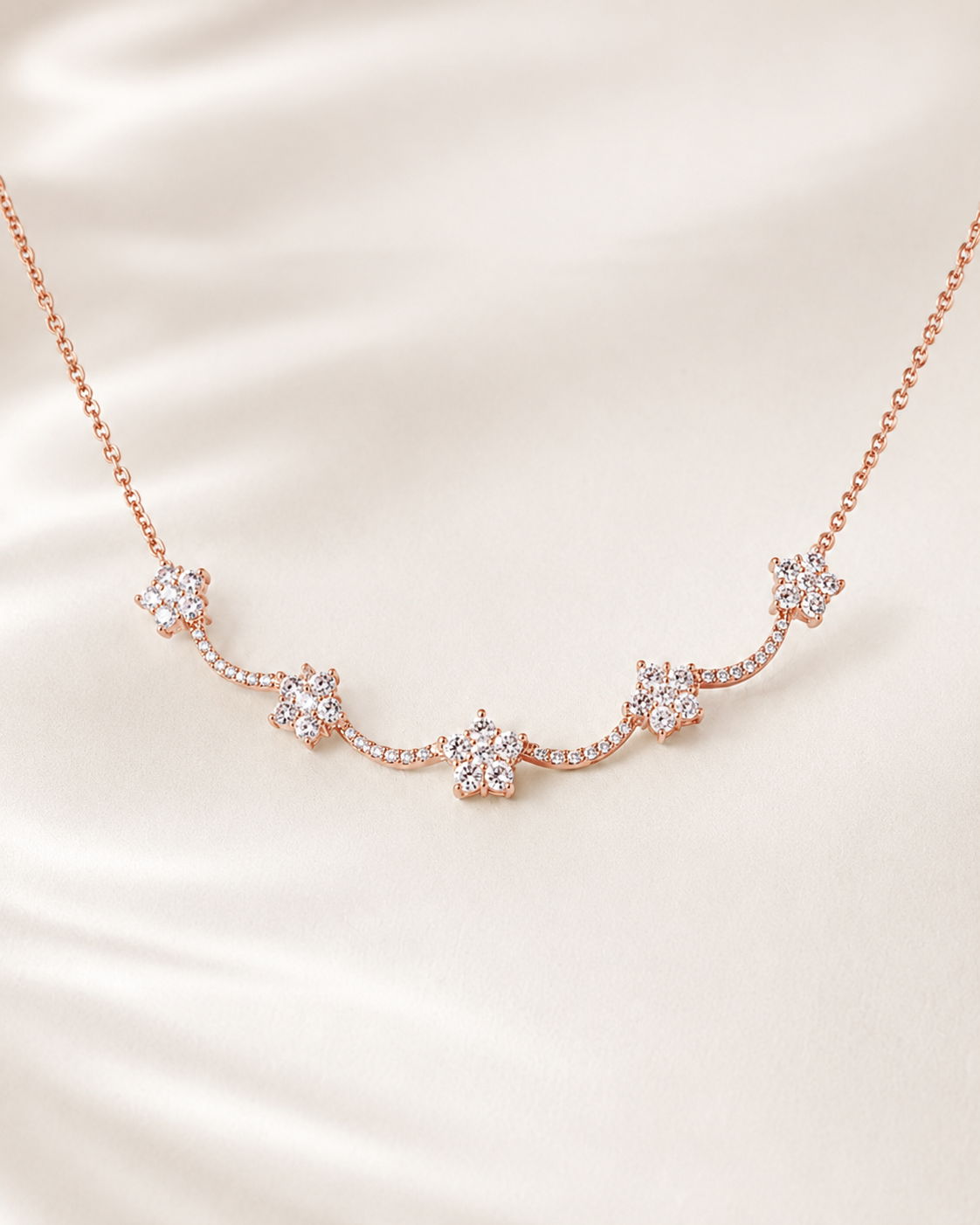 A delicate rose gold necklace featuring five small star-shaped clusters of clear crystals, displayed on a soft, cream-colored satin fabric background.