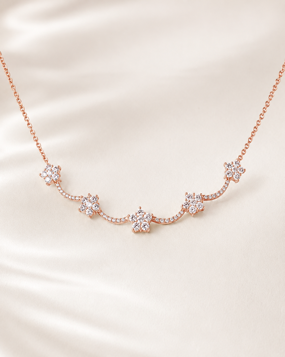 A delicate rose gold necklace featuring five small star-shaped clusters of clear crystals, displayed on a soft, cream-colored satin fabric background.