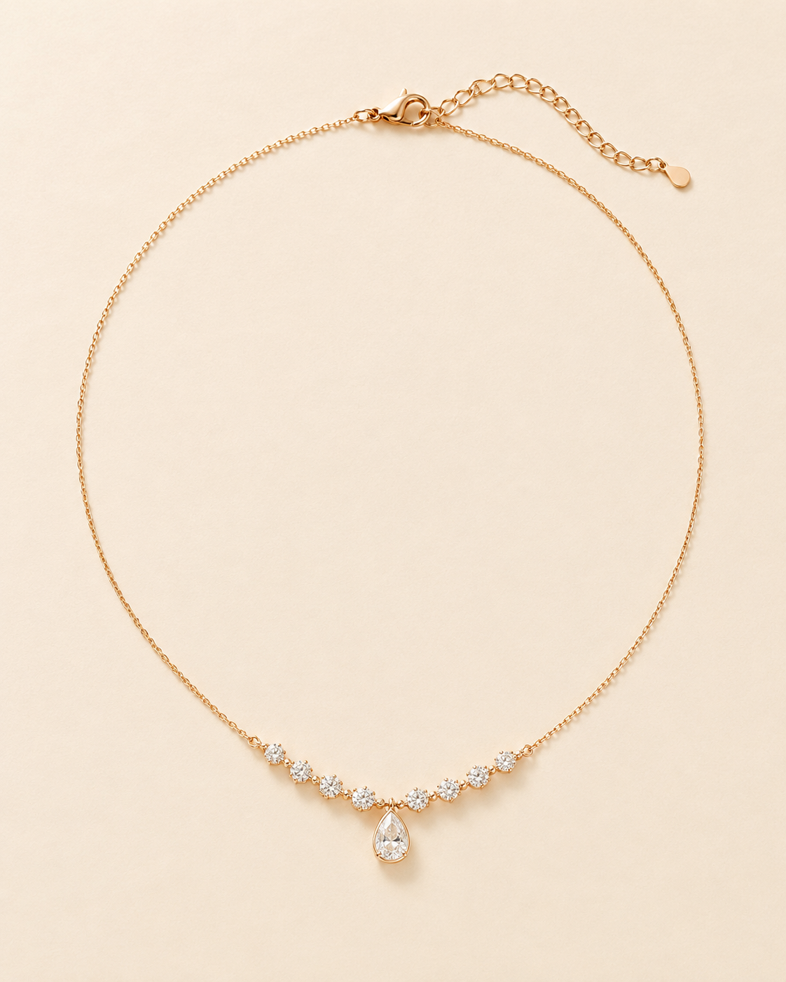 A delicate gold chain necklace with a curved row of small clear stones and a teardrop-shaped clear stone pendant, displayed on a light beige background.