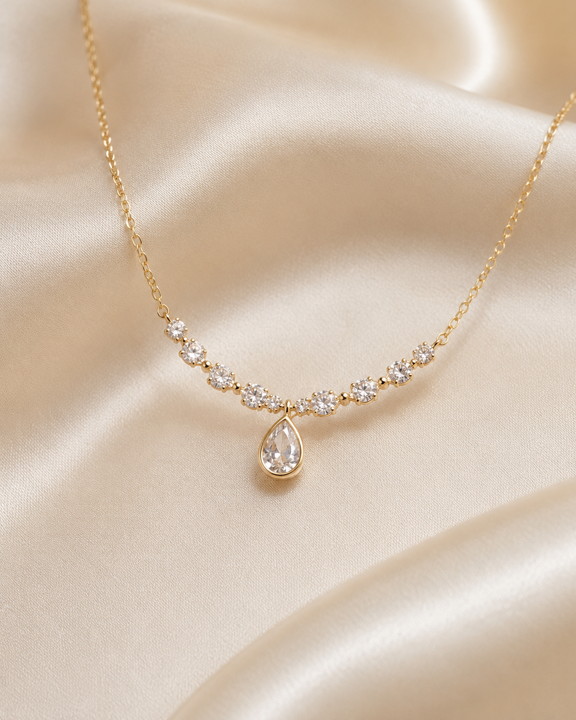 A delicate gold necklace with a curved row of small clear stones and a teardrop-shaped clear stone pendant, displayed on draped champagne satin fabric.