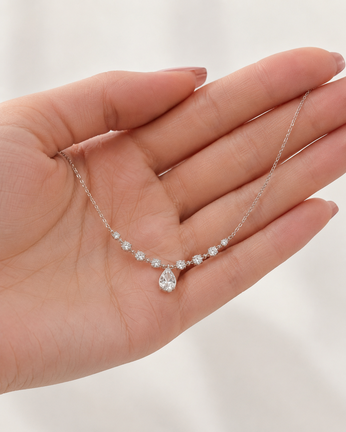 A delicate silver necklace with a pear-cut center stone and small clear accent stones, displayed on an open hand against a soft white background.