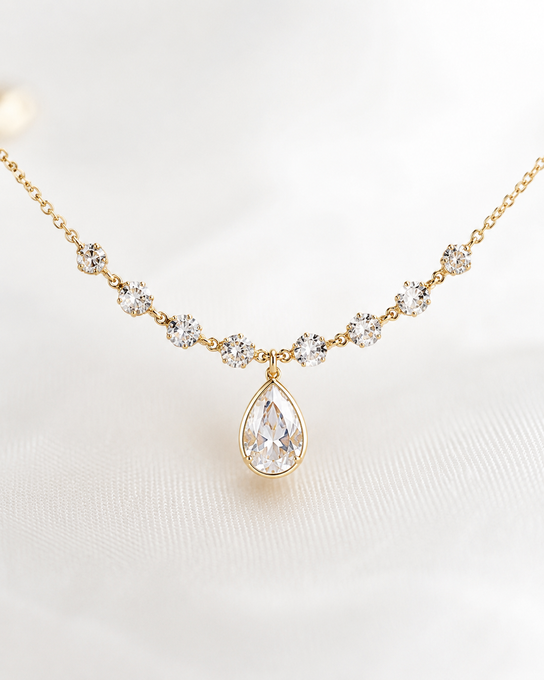 A gold necklace featuring a teardrop-shaped clear gemstone pendant and smaller clear gemstone floral accents along the chain, displayed on a soft white fabric background.