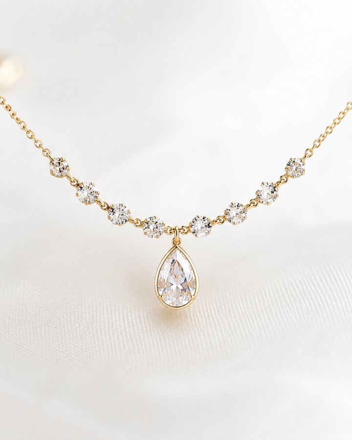 A gold necklace featuring a teardrop-shaped clear gemstone pendant and smaller clear gemstone floral accents along the chain, displayed on a soft white fabric background.