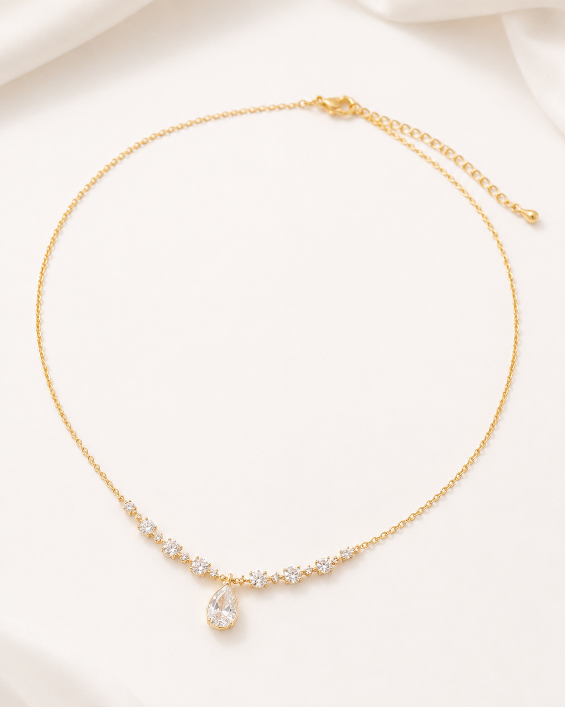 A delicate gold chain necklace with a small row of clear stones and a teardrop-shaped clear stone pendant, displayed on a soft white fabric background.