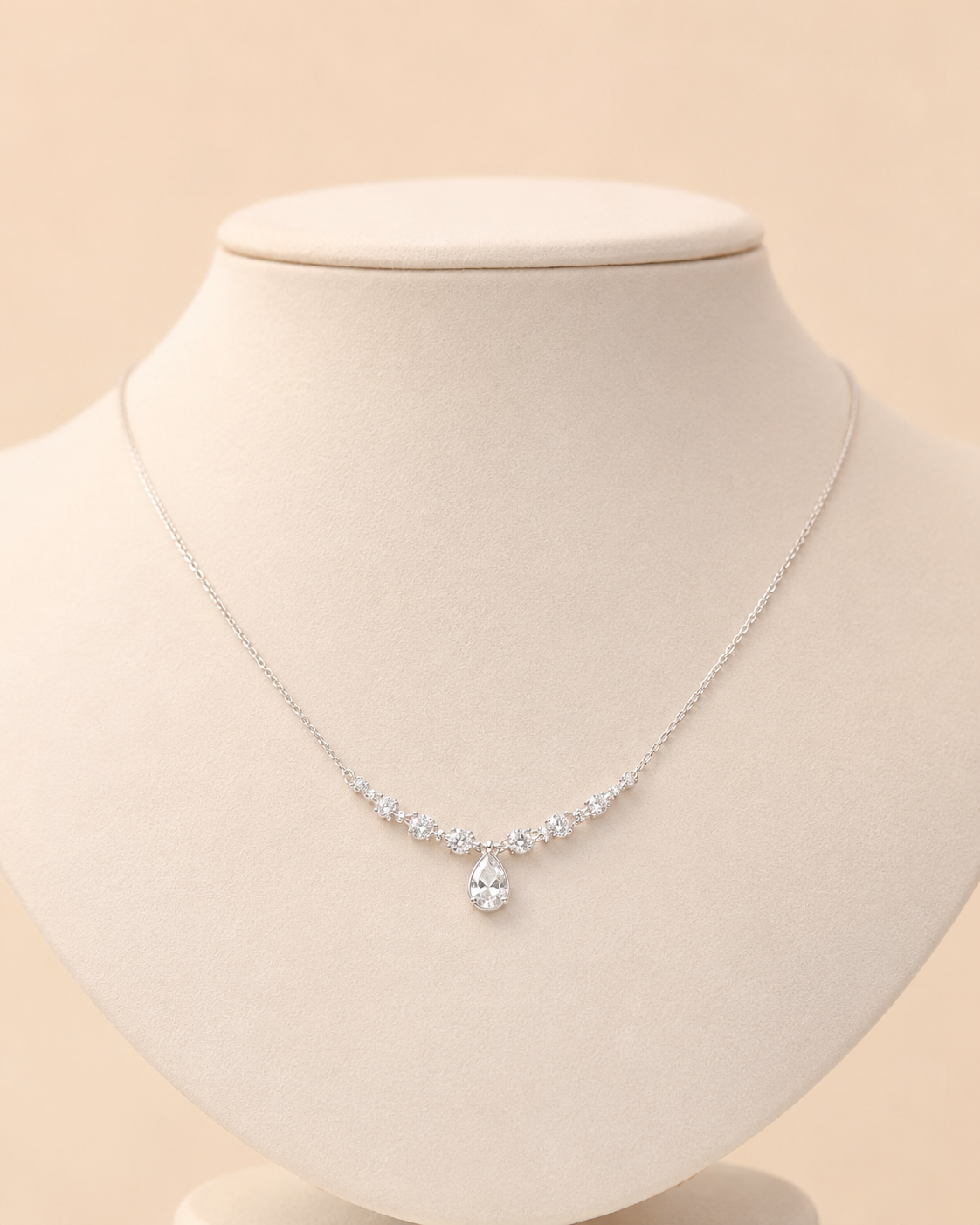 A delicate silver necklace with a small cluster of clear stones and a pear-shaped clear center stone, displayed on a cream-colored jewelry bust against a soft beige background.