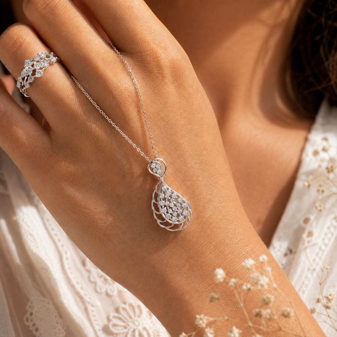 A close-up lifestyle shot of a woman's hand wearing a delicate silver teardrop pendant necklace and a matching silver ring with clear stones, styled with a white lace top.