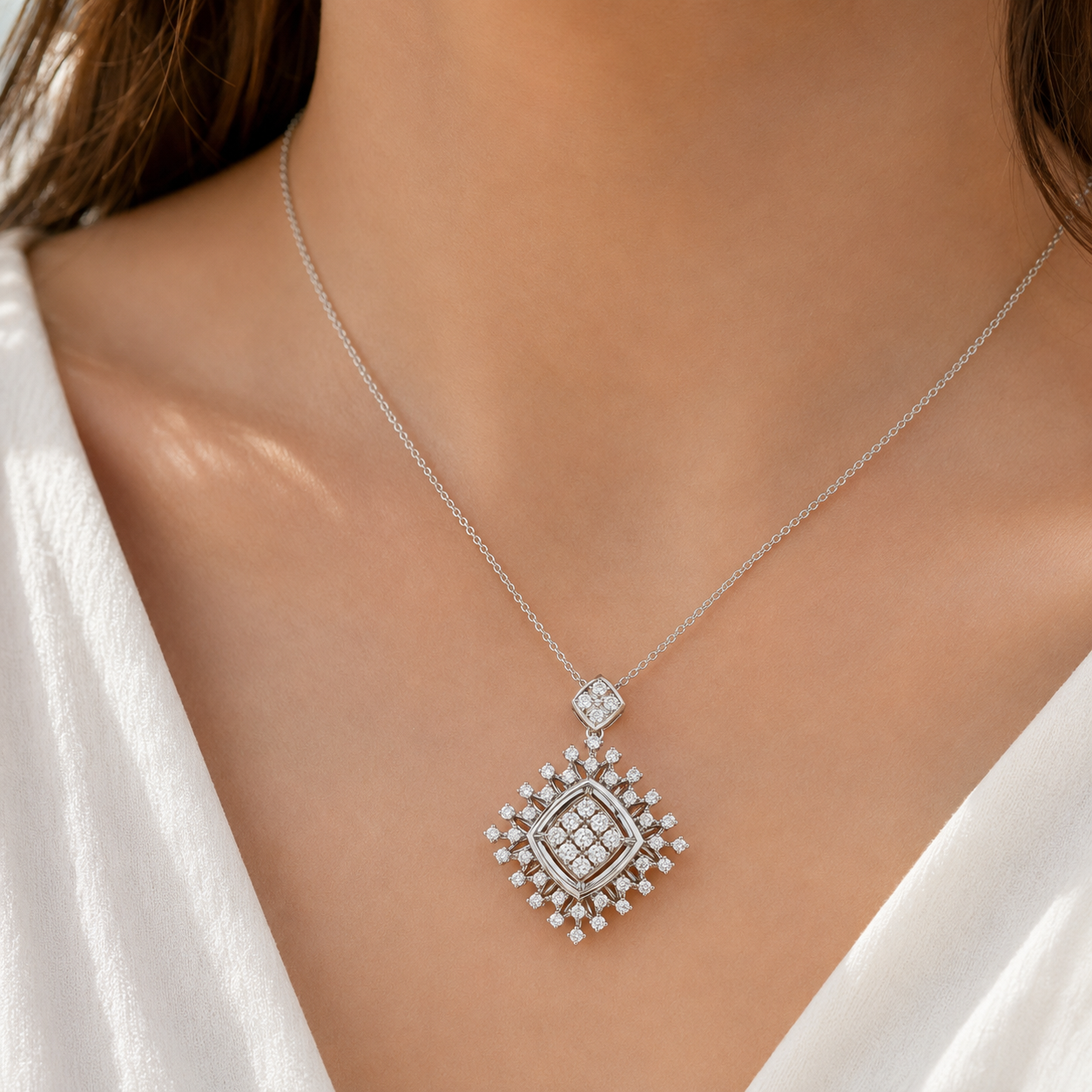 A close-up of a model wearing a delicate silver chain necklace with a diamond-encrusted pendant. The pendant features a central square cluster of clear stones surrounded by a halo of smaller stones, suspended from a fine silver chain.