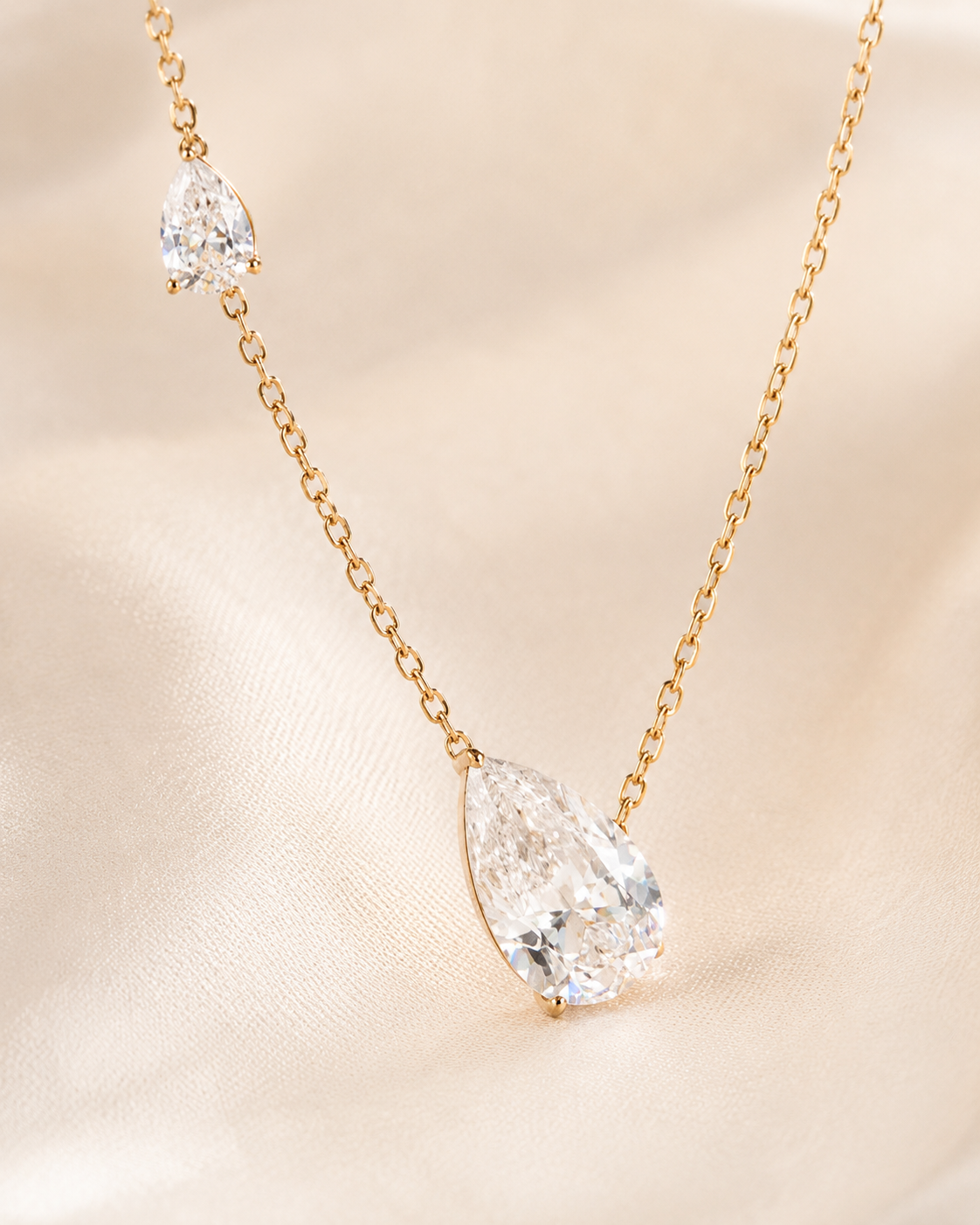 A delicate gold chain necklace featuring a pear-cut clear gemstone pendant and a smaller matching gemstone accent on the chain, displayed on a soft beige satin fabric background.