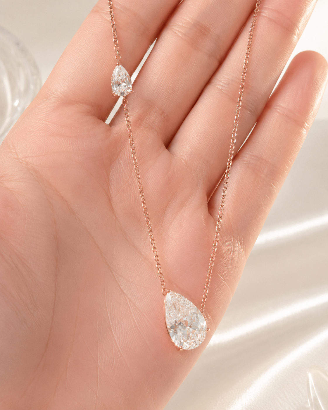 A delicate rose gold necklace with a large teardrop-shaped clear gemstone pendant, held in the palm of a hand.