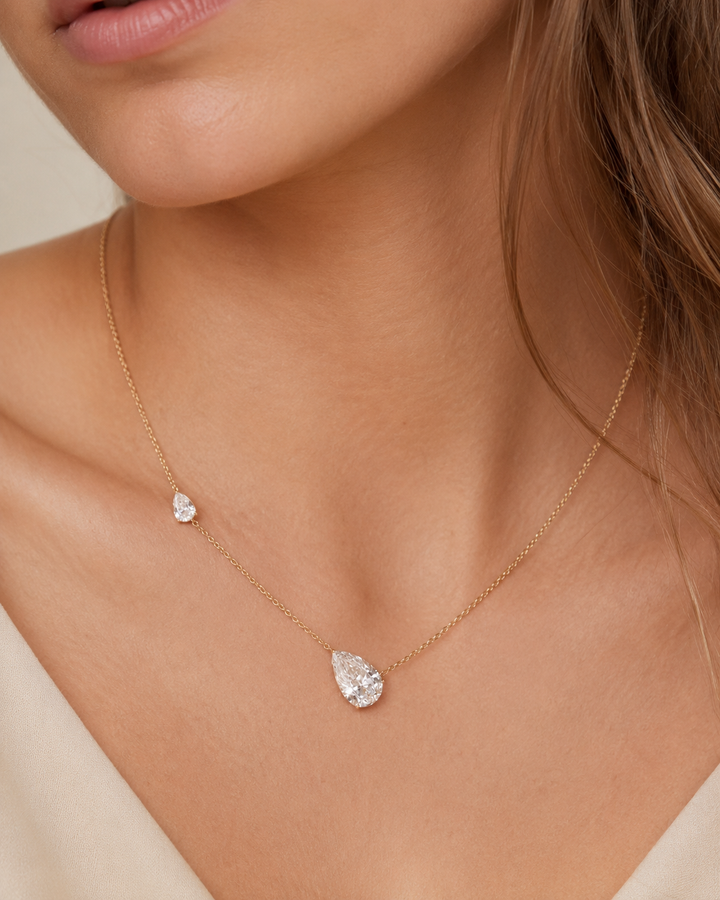 A close-up of a model wearing a delicate gold chain necklace with two clear teardrop-shaped gemstones, one larger and one smaller.