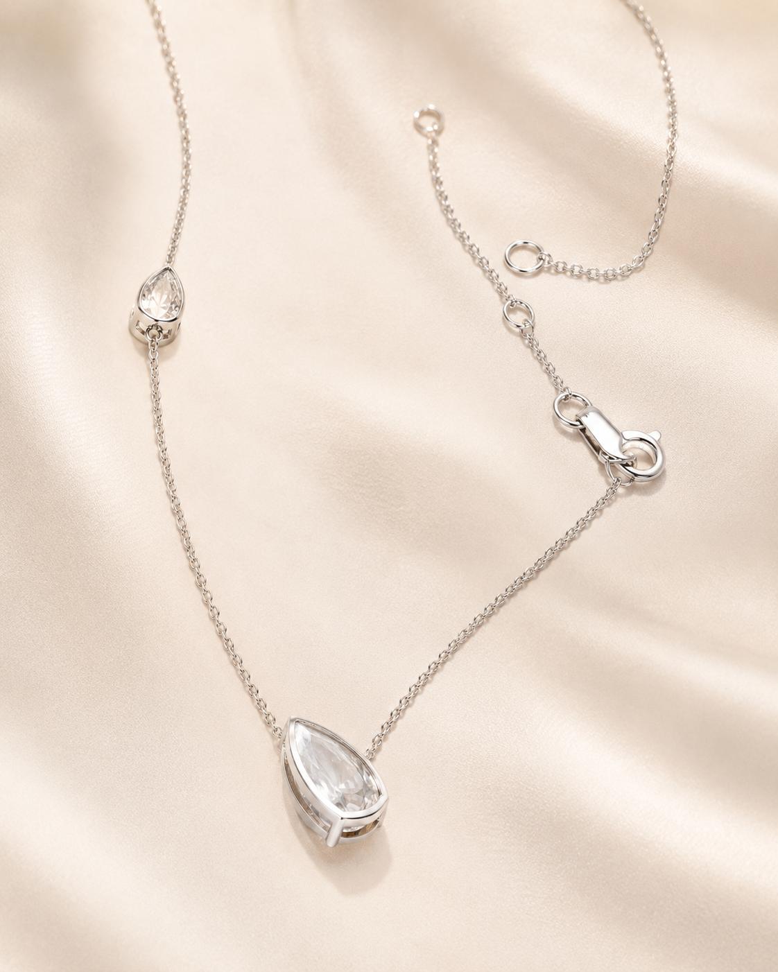 A delicate silver chain necklace with a teardrop-shaped clear gemstone pendant, displayed on a soft beige satin fabric background.