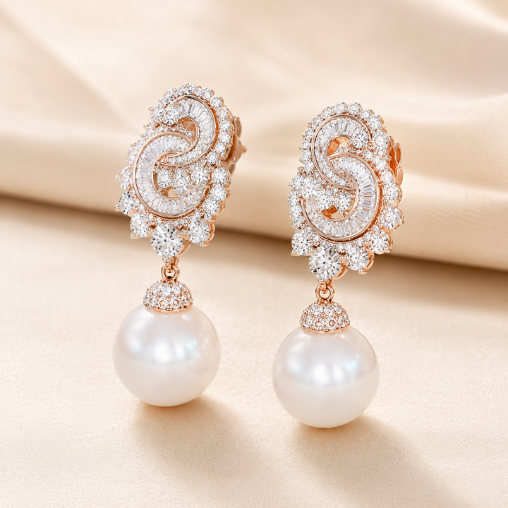 A pair of rose gold drop earrings featuring intricate pavé-set clear stones in a swirling design, with a large white pearl hanging from each earring, displayed on a soft beige satin fabric background.