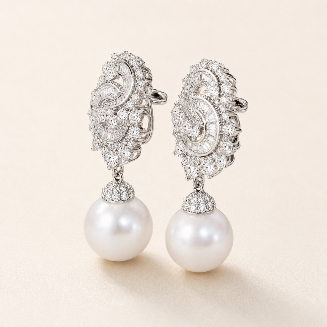 A pair of ornate silver drop earrings featuring intricate pavé-set clear stones and a dangling white pearl, shown on a light beige background.