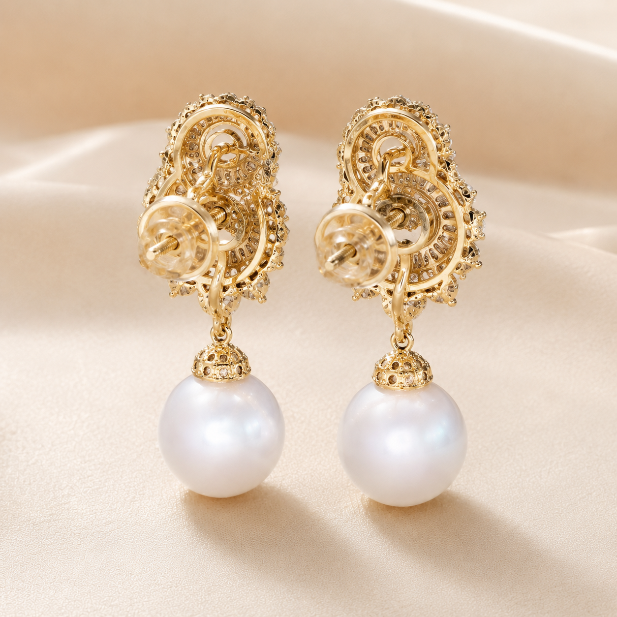 A pair of ornate gold drop earrings with intricate filigree detailing and a large white pearl hanging from each earring, displayed on a soft beige fabric background.