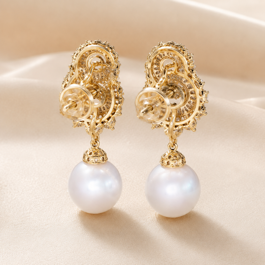 A pair of ornate gold drop earrings with intricate filigree detailing and a large white pearl hanging from each earring, displayed on a soft beige fabric background.