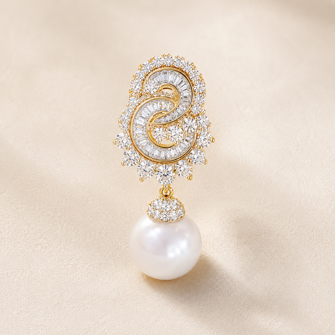 A single gold and crystal drop earring featuring a circular pavé crystal design with a central gold swirl and a dangling white pearl, set against a soft beige background.
