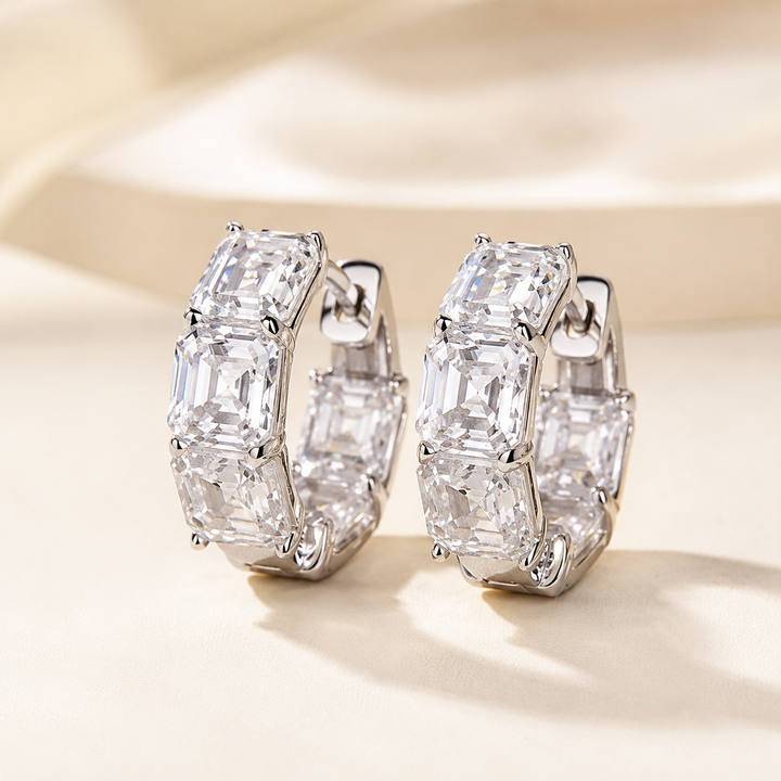 A pair of silver hoop earrings with large, clear baguette-cut gemstones, displayed on a soft beige fabric background.