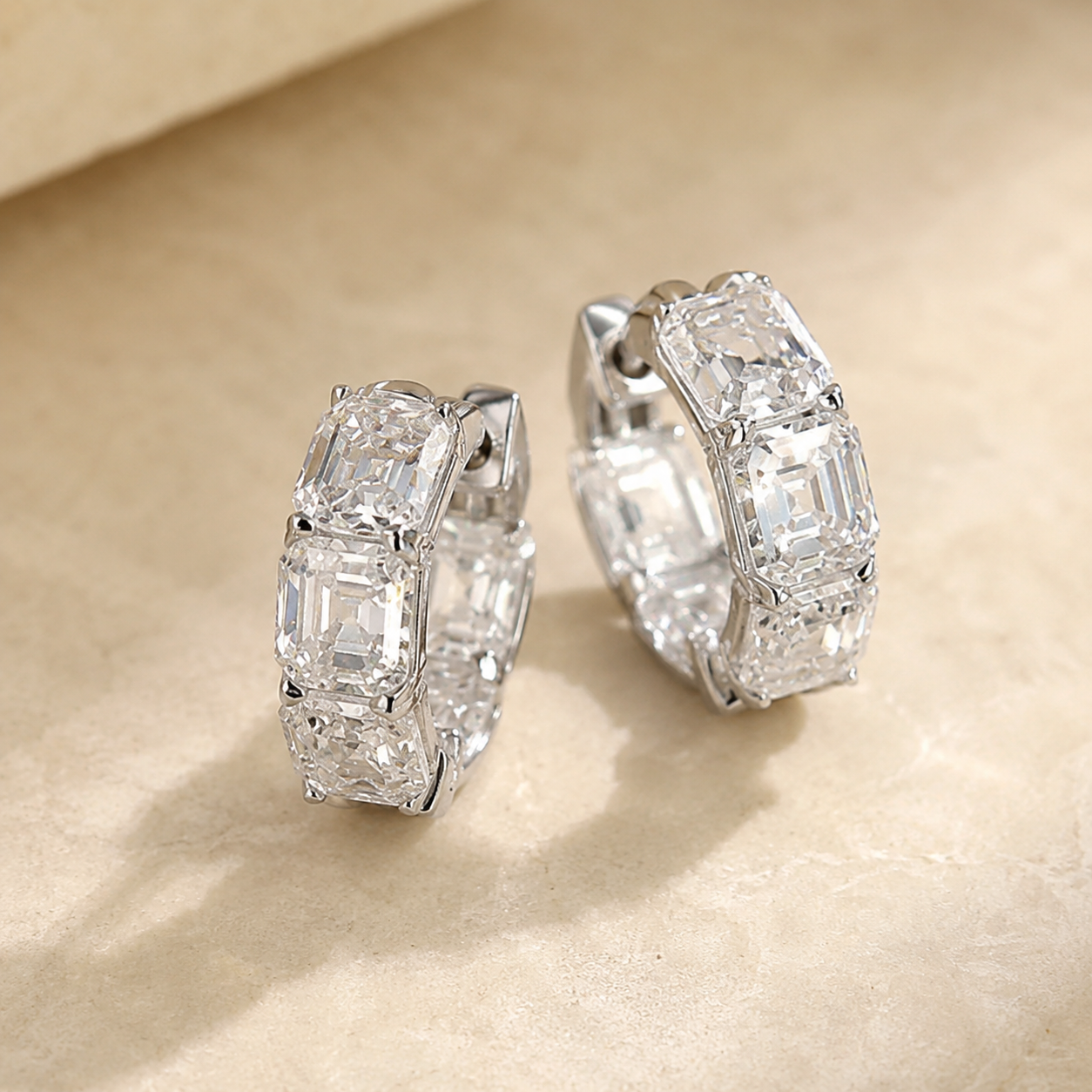A pair of silver hoop earrings set with clear baguette-cut gemstones, displayed on a textured beige surface.
