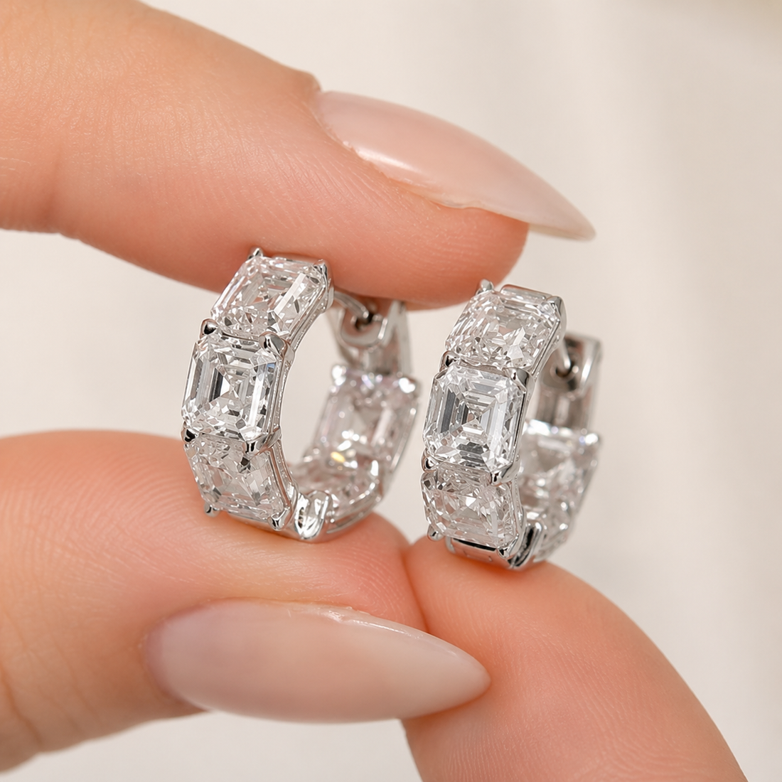 A close-up of a person's hand with manicured nails holding a pair of small silver huggie hoop earrings set with clear baguette and round-cut gemstones against a soft, light beige background.