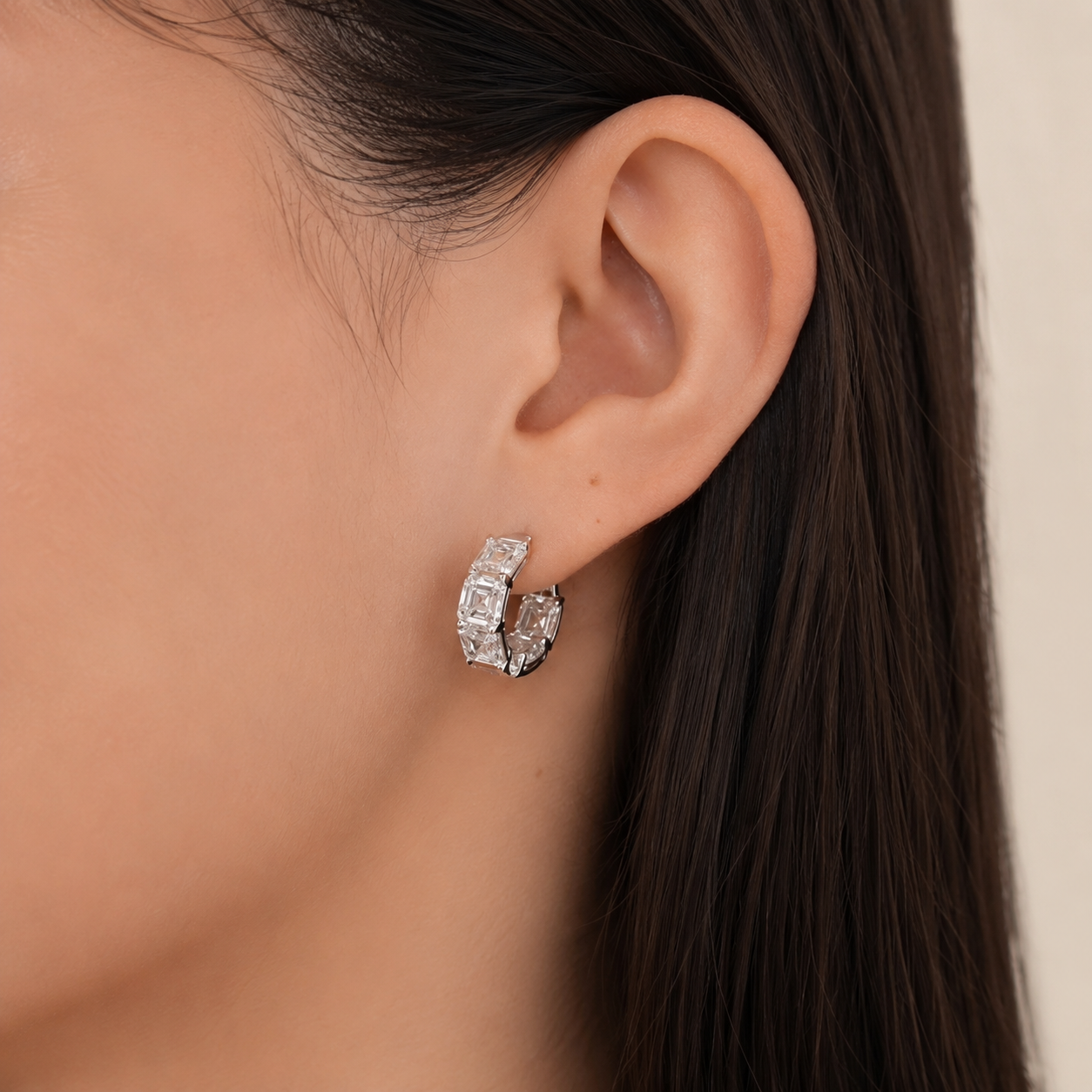 A close-up of a model's ear wearing a small silver huggie hoop earring adorned with clear rectangular crystals.