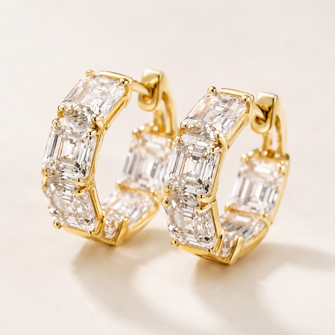 A pair of gold hoop earrings set with large clear baguette-cut diamonds, photographed on a clean light beige background.